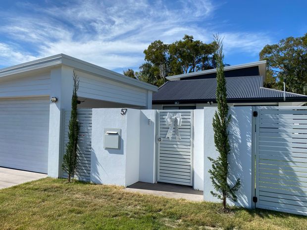Street-facing view of a contemporary single-storey residence with a white rendered and horizontal-clad facade, attached garage, pedestrian entry gate, slatted front fencing, dark pitched metal roof, and narrow columnar landscaping.