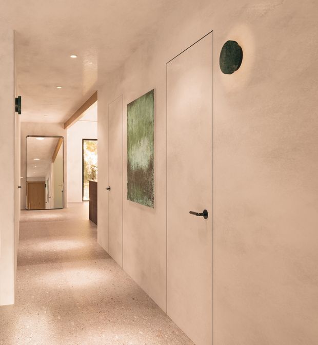 Warm, minimalist interior hallway with seamless pale plastered walls, flush concealed doors, dark minimalist hardware, and a terrazzo-like speckled floor. A large mirror reflects the corridor, while a narrow window brings in natural light. The space has a refined, softly textured and monochromatic finish.