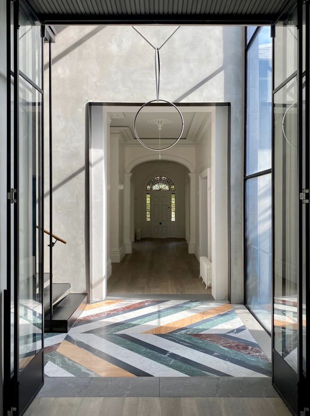 A formal residential entry sequence viewed through open black-framed glass doors. The foreground features a striking geometric patterned stone floor with diagonal bands in black, white, green, terracotta, and wood tones. Beyond it is a high-ceilinged hallway with classic white architectural mouldings, an arched opening, dark timber flooring, a central pendant light, and a glazed-panel front door. A stair with a timber handrail is visible at left, while strong daylight casts angular shadows across the walls.