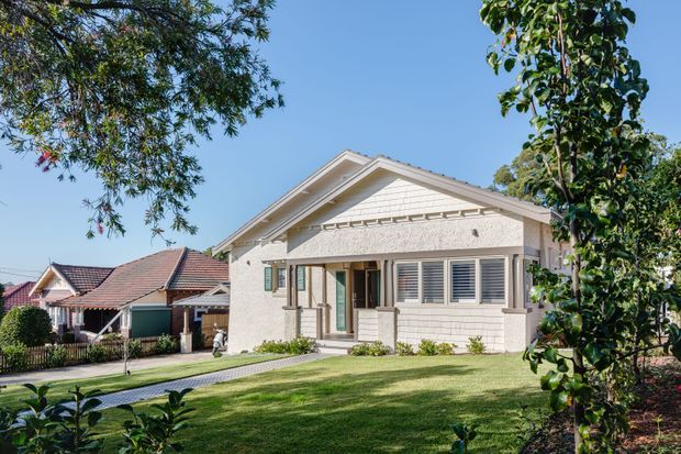 Front exterior view of a single-storey suburban house with a prominent gabled roof, pale rendered and shingle-like facade detailing, wide louvred windows, and a covered entry porch. The neatly landscaped front lawn, paved path, mature planting, and neighbouring pitched-roof homes are visible. The architecture has restrained traditional and bungalow-influenced detailing.