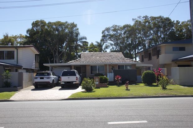 Street-facing view of a single-storey suburban house with a low tiled hip roof, pale rendered walls, a covered front entry, front lawn, driveway and two parked vehicles. Mature trees and neighbouring homes surround the property.
