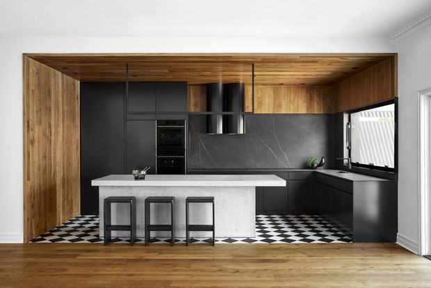 A contemporary kitchen with a dramatic black-and-white checkerboard floor, matte black cabinetry, dark stone-look splashback, and a large light stone island with three stools. Warm timber wall and ceiling lining frames the kitchen, while black-framed glazing brings in light from the right.