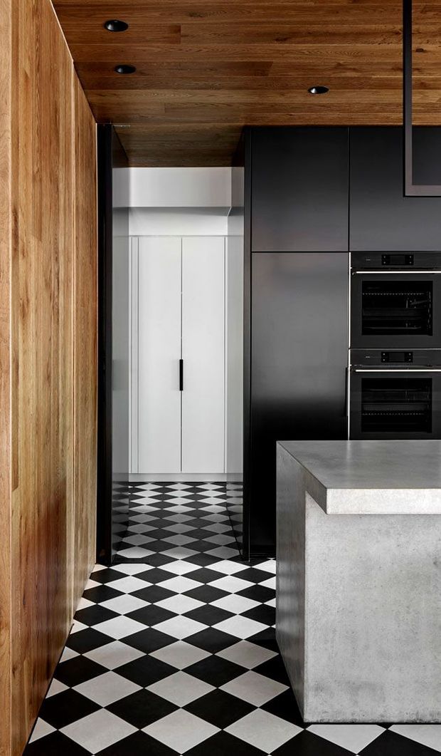 A compact, highly designed modern kitchen featuring dark matte cabinetry with integrated double ovens, a substantial grey concrete island, rich timber wall and ceiling lining, and a dramatic black-and-white checkerboard tiled floor. A narrow white pantry or storage passage is visible beyond the kitchen.