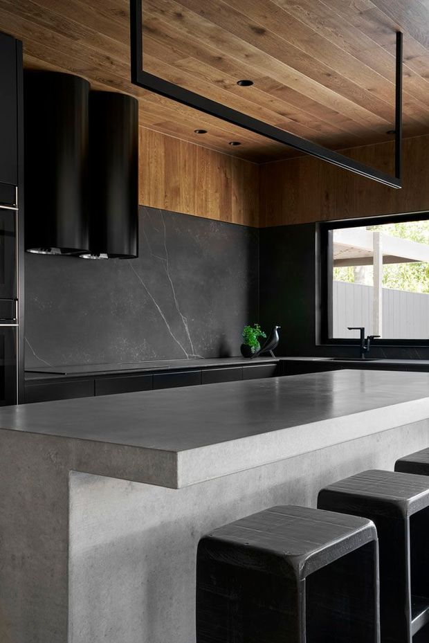 A dramatic contemporary kitchen with a large concrete island and dark stone benchtop, matte-black cabinetry, black rangehoods and fixtures, and warm timber-lined ceiling and upper walls. Black upholstered bar stools sit at the island, while a broad window brings in natural light.