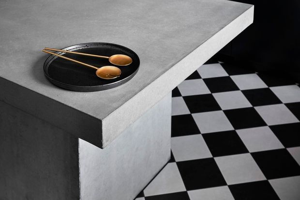 Close-up architectural detail of a minimalist concrete island or counter with a smooth grey slab and monolithic base. A black dish with wooden utensils sits on the surface, while a bold black-and-white checkerboard floor extends into the background.
