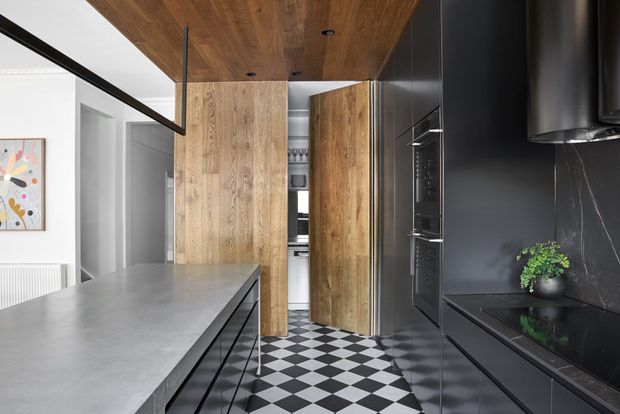 A contemporary galley kitchen with a large grey island, matte black cabinetry and appliances, warm timber ceiling and tall timber pantry doors. The space features a dramatic black-and-white checkerboard floor, dark stone backsplash, integrated lighting, and a glimpse of an adjacent white-walled area.