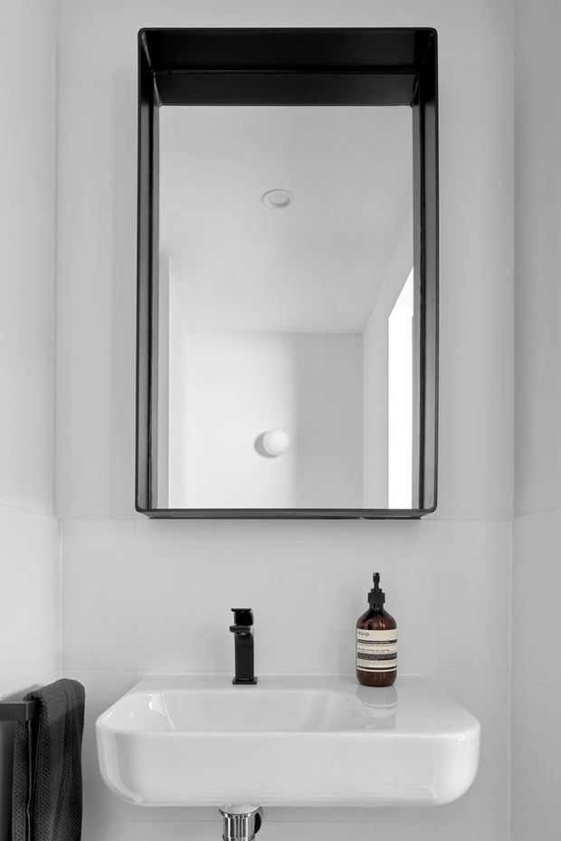 A compact contemporary bathroom vanity with a white wall-mounted basin, black tapware, dark-framed rectangular mirror, white tiled walls, and a small amber soap dispenser. A dark towel hangs at the side.