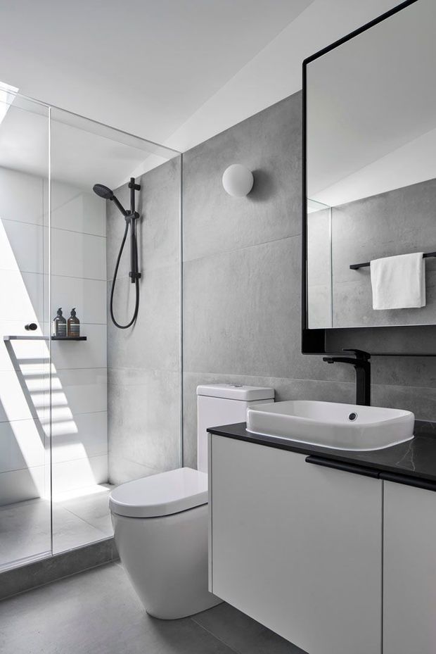 A contemporary bathroom with a frameless glass walk-in shower, large grey concrete-look wall tiles, a wall-mounted round light, white toilet, and a floating vanity with a white vessel basin and dark benchtop. A large black-framed mirror and towel rail sit above the vanity. Bright natural light creates strong linear shadows across the shower floor.