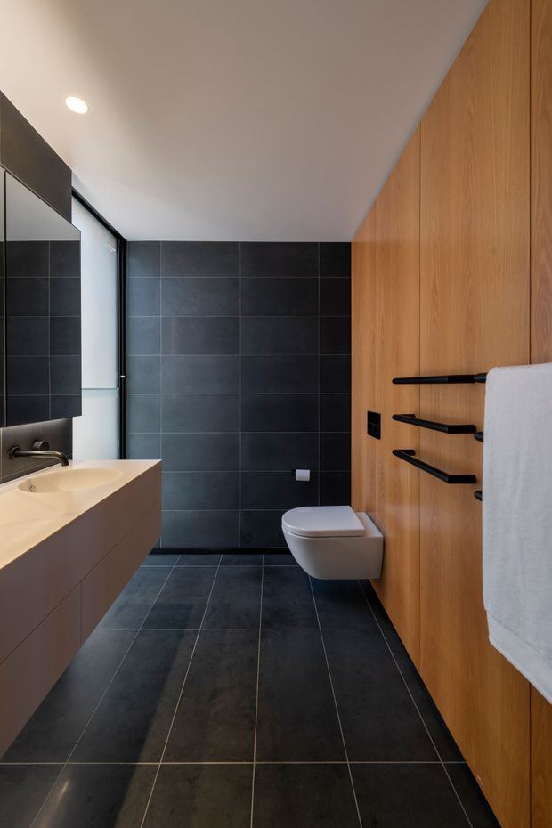 Contemporary bathroom with dark large-format tiled walls and floor, a floating light-toned vanity with integrated basin, wall-mounted toilet, black tapware and accessories, and full-height warm timber wall cabinetry or panelling. A frameless glass shower enclosure is visible at the rear, with clean minimalist detailing and recessed ceiling lighting.