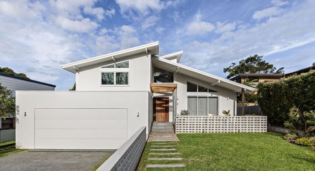 Street-facing exterior of a contemporary single-storey residence with a strong asymmetrical sloping roofline, white vertical cladding, an integrated double garage, timber-accented entry, and a patterned breeze-block privacy wall. The front garden, stepping-stone path, and open lawn are visible under a partly cloudy sky.