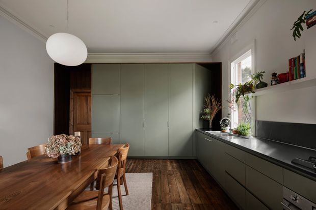 A warm, refined kitchen-dining interior with a long walnut dining table and timber chairs in the foreground. The kitchen features full-height muted sage-green cabinetry, dark benchtops, a black splashback, open white shelving, and a large window with plants. Dark timber flooring, detailed cornices, and a spherical pendant light add character to the otherwise streamlined space.