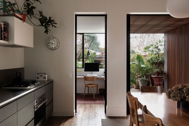 A warm, contemporary open-plan kitchen viewed toward a compact home office and a plant-filled adjoining space. The kitchen features dark cabinetry, a pale benchtop, integrated appliances, open shelving, and a small breakfast or dining area. Timber flooring continues into the office, which has a desk, chair, rug, and large black-framed window. The adjoining space includes timber wall and ceiling slats, abundant indoor plants, and a dining table with timber chairs. A large spherical pendant light is partially visible overhead.