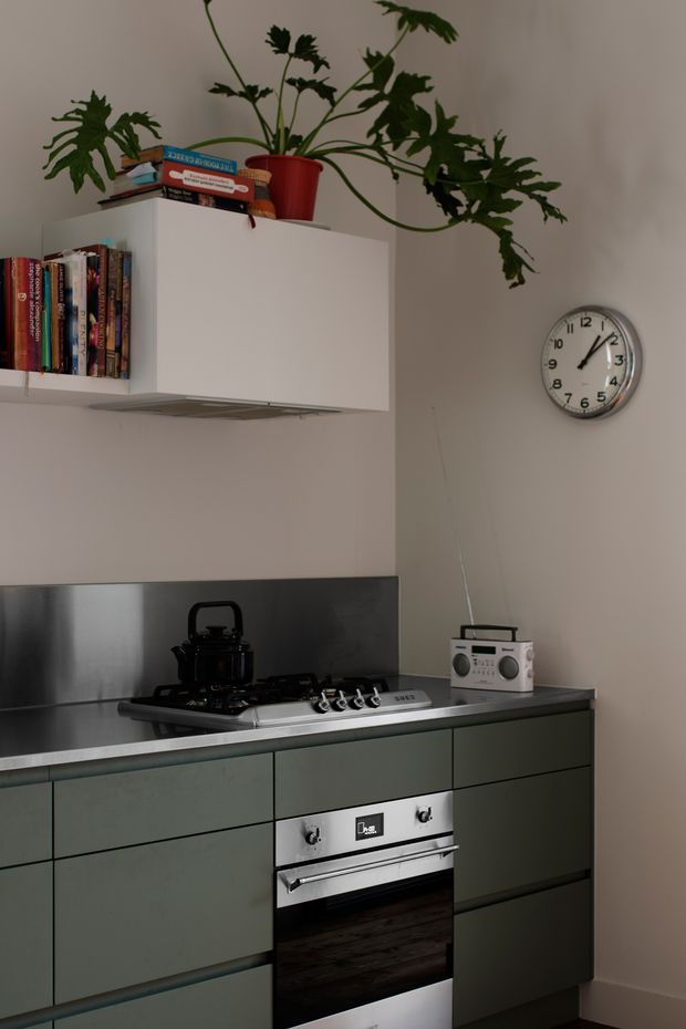 A compact modern kitchen with muted green flat-panel cabinetry, a dark benchtop, stainless-steel splashback, and an integrated stainless-steel oven and cooktop. White wall-mounted cabinetry includes an open bookshelf section, while a large trailing indoor plant sits on top. A wall clock and small radio add everyday character to the softly lit space.