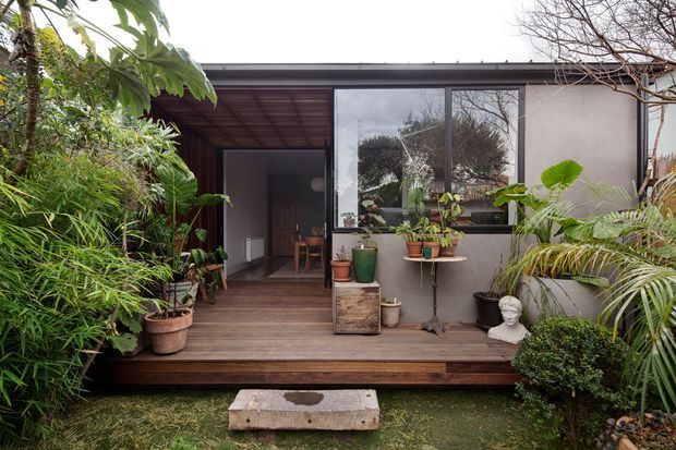 A compact modern dwelling viewed from a lush garden courtyard. The facade has a dark low-pitched roof edge, smooth light-grey wall finish, large dark-framed glazing, and a recessed covered entry opening onto a raised timber deck. Numerous potted tropical and leafy plants surround the deck, with a simple concrete stepping stone in the foreground. The open doorway reveals a small interior seating area.