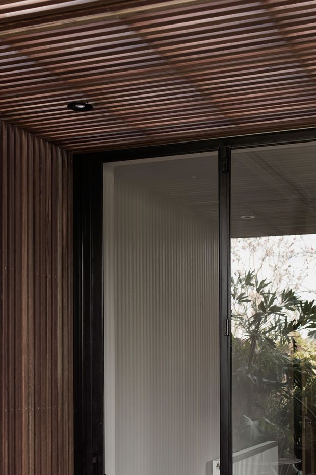 Close-up architectural detail of a dark timber-lined exterior alcove or covered entry, with finely spaced slatted timber across the ceiling and side wall. A black-framed glazed door or window occupies the right side, reflecting surrounding vegetation and revealing a dim interior beyond. A recessed circular downlight is integrated into the timber ceiling. The composition emphasizes warm dark wood texture, black framing, and a contemporary sheltered threshold.
