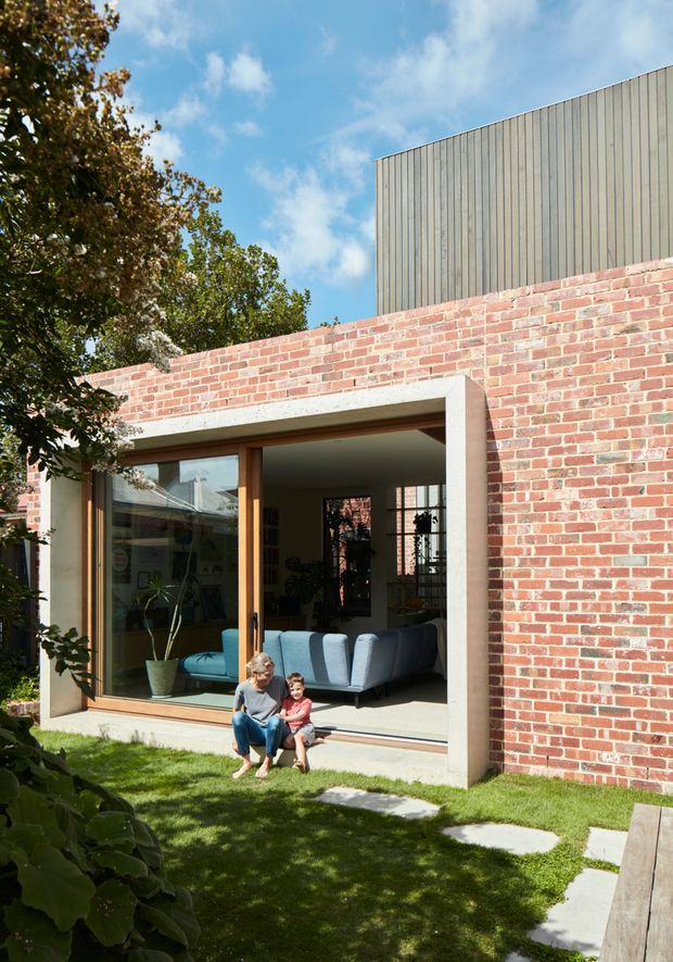 Exterior view of a contemporary brick house opening onto a small landscaped courtyard or lawn. The facade combines exposed red brickwork with a tall vertically clad upper volume and a large timber-framed glazed opening. A living room with a blue sofa is visible inside, while stepping stones, lawn, mature planting, and people provide scale.