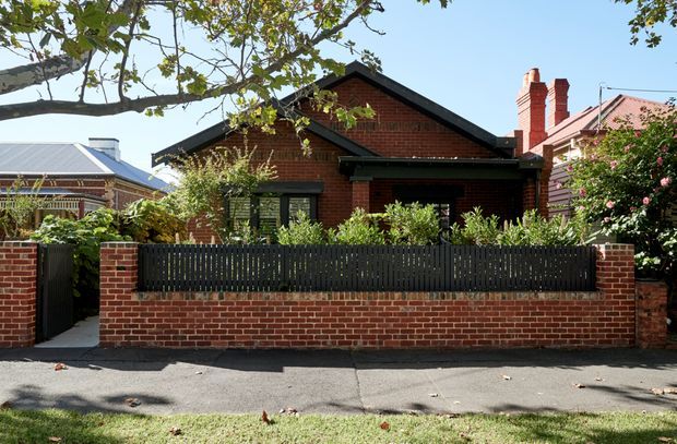 Street-facing view of a single-storey red-brick house with a prominent dark gabled roof, black-framed openings, and a brick boundary wall topped with dark vertical fencing. Mature trees and garden planting frame the suburban residential facade, with neighbouring period-style homes visible on either side.