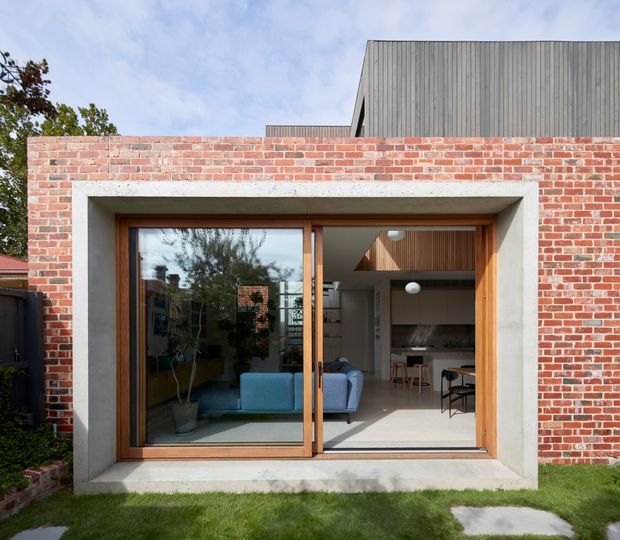 Exterior view of a contemporary brick dwelling with a large timber-framed glazed sliding door opening into a bright living area and kitchen. The facade combines red brickwork, a deep pale concrete surround, timber joinery, and an upper vertical cladding volume. A sofa, dining area, kitchen cabinetry, and pendant lights are visible inside through the glazing.