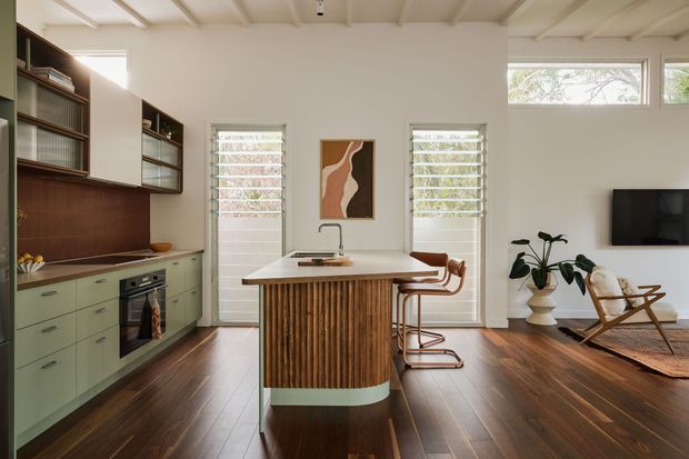 A bright open-plan kitchen and living area with a central island featuring vertical timber slats, pale green cabinetry, a dark timber splashback, and a light benchtop. The room has dark timber flooring, white walls, exposed painted ceiling beams, plantation-shuttered windows and doors, a dining nook, and a small living area with a wall-mounted television and lounge chairs. The overall design is warm, refined, and contemporary with mid-century influences.