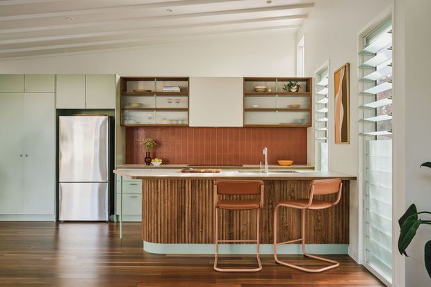 A bright, compact contemporary kitchen with a central island and integrated seating. The room features pale flat-panel cabinetry, a stainless-steel refrigerator, open timber shelving, a terracotta-toned vertical tile splashback, and a distinctive fluted timber island front. White painted walls and a coffered or battened ceiling create an airy setting, while louvred windows and doors provide ventilation and daylight. Two timber-and-metal bar stools sit at the island, and dark timber flooring adds warmth.