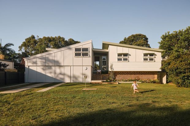 A suburban single-storey house viewed from the front, featuring a pale rendered or painted facade, low-pitched asymmetrical roof forms, horizontal windows, a recessed entry, and a raised section supported by a decorative timber lattice base. A broad grass lawn and mature trees surround the home.