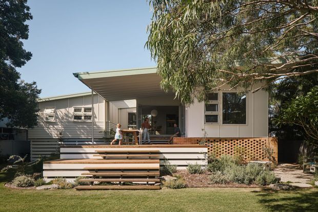A single-storey residential house viewed from the garden, featuring a low-pitched roof, light horizontal weatherboard cladding, and a broad covered outdoor deck. Wide tiered timber steps lead up through a landscaped front or rear garden to the deck, with brick latticework beneath the platform and large windows opening onto the outdoor living area. Mature trees partially frame the home.