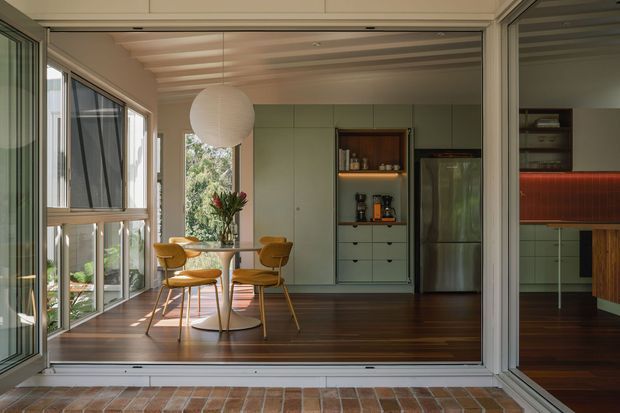 A bright, mid-century-inspired kitchen and dining area viewed from a covered outdoor threshold through fully glazed sliding doors. The room features dark timber flooring, pale green built-in cabinetry, stainless-steel refrigeration, a warm red tiled splashback visible to the right, and a compact round dining table with mustard upholstered chairs. A large globe pendant, extensive windows, and a partially louvred ceiling create a warm indoor-outdoor connection.