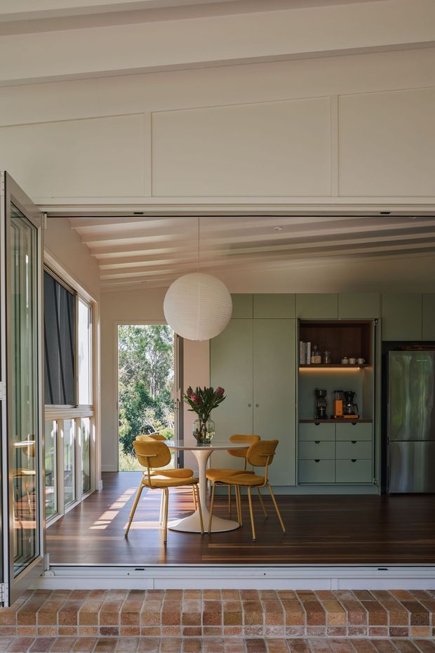A warm, carefully detailed dining and kitchen space viewed through a wide open glazed opening. The room features dark timber flooring, pale green built-in cabinetry, a concealed appliance wall with open shelving, a round white dining table, mustard upholstered chairs, and a large spherical pendant light. Timber-framed glazing opens to leafy surroundings, while a brick threshold and broad masonry step connect the interior to the exterior. Exposed ceiling battens add architectural texture and rhythm.