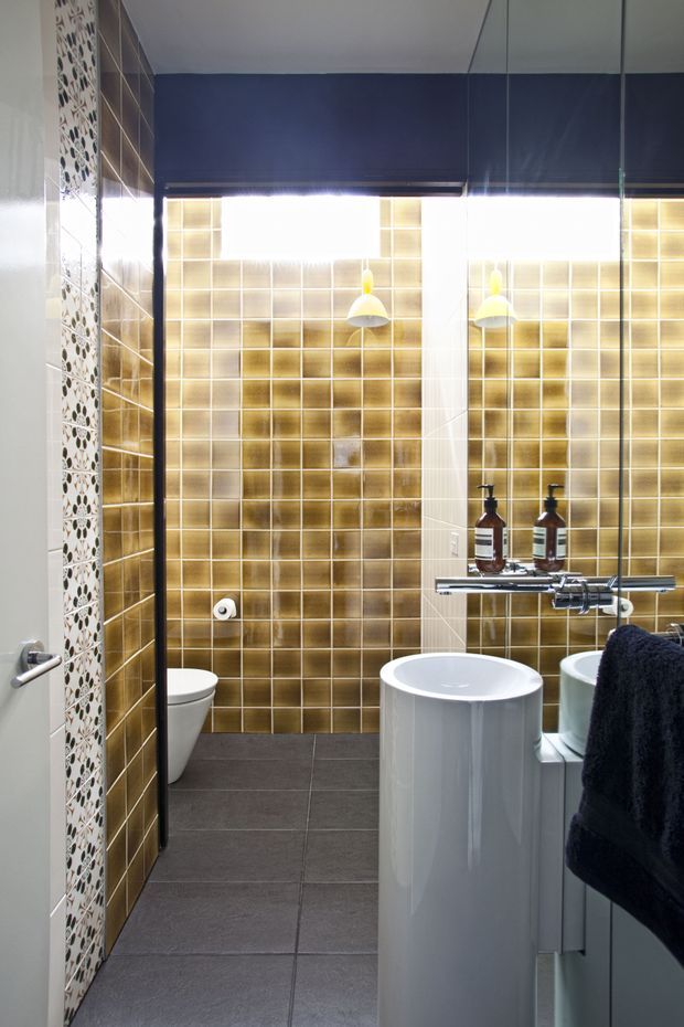 Compact contemporary bathroom with a freestanding white basin, wall-mounted chrome tapware, toilet, dark grey floor tiles, and distinctive glossy gold-toned square wall tiles. A mirrored cabinet reflects the basin and fixtures, while a dark navy roller blind and patterned vertical tile strip add contrast.