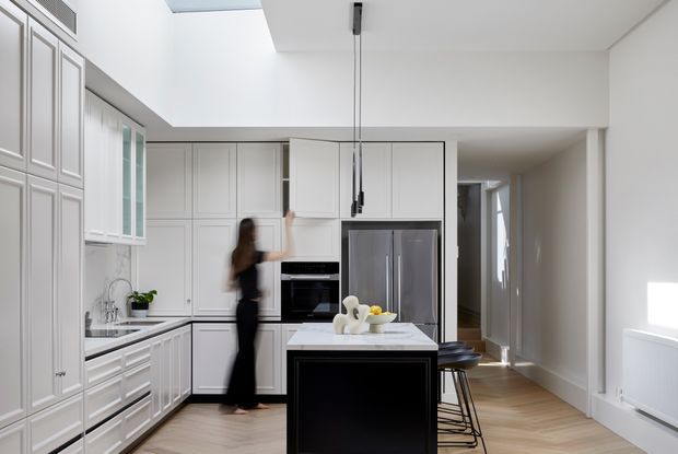 Bright contemporary kitchen with white framed cabinetry, stainless-steel appliances, a dark island base with a pale stone benchtop, and timber flooring. A skylight brings natural light into the open-plan space, with a hallway visible beyond.