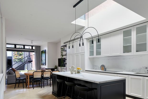 Bright open-plan kitchen and dining area with extensive white shaker-style cabinetry, a dark island with seating, pale timber flooring, marble-look splashback, and sculptural black pendant lights. A dining table and living area extend toward large glazed doors at the rear, while a skylight brings additional natural light into the kitchen.