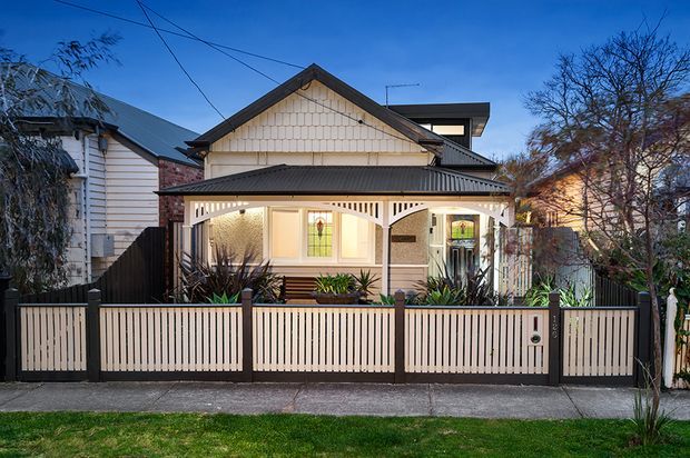 Street-facing view of a narrow traditional home with a prominent front gable, decorative shingle cladding, covered veranda, timber detailing, and a contemporary rear roof extension. The property has a low front fence and landscaped garden beds, photographed at dusk.