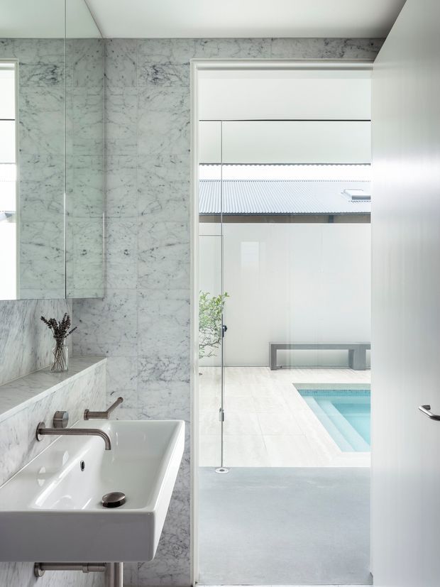 Bright contemporary bathroom featuring a wall-mounted white rectangular basin with brushed metal tapware, a freestanding white bathtub, and extensive pale veined marble wall cladding. A full-height glazed opening provides a view toward a private courtyard and turquoise swimming pool. The space is clean-lined, highly reflective, and filled with natural light.