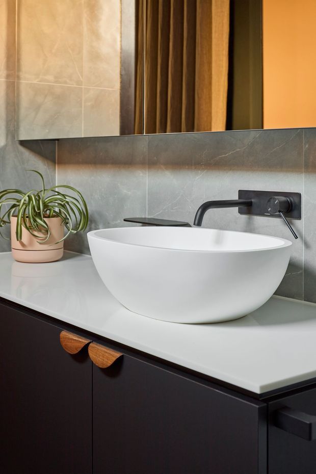 A close-up contemporary bathroom vanity featuring a white vessel basin on a pale stone-look benchtop, a matte black wall-mounted tap, dark cabinetry with timber handles, grey stone-look tiled splashback, and a large mirrored cabinet. A potted spider plant adds a small natural accent.