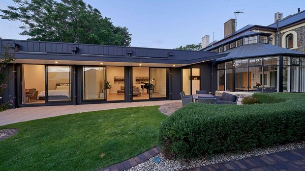 Exterior view of a landscaped residential courtyard showing a dark contemporary single-storey extension with a low flat roof, extensive floor-to-ceiling glazing, and recessed outdoor lighting. The extension connects to a larger traditional multi-storey house with pitched roof elements, masonry walls, arched windows, and chimneys. A paved patio with outdoor seating, manicured hedges, lawn, and garden edging occupies the foreground.