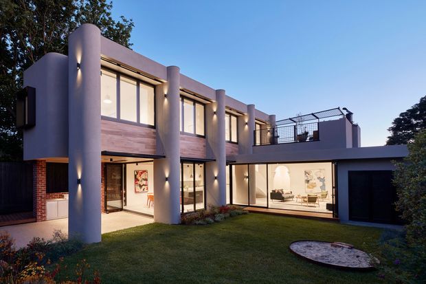 Twilight exterior view of a contemporary multi-storey residence arranged around a landscaped lawn courtyard. The facade combines smooth rounded vertical columns, extensive floor-to-ceiling glazing, warm timber-look wall panels, dark brick sections, and a flat roofline. Illuminated interiors are visible through the windows, while an upper terrace with metal balustrading and a dark garage opening complete the composition.