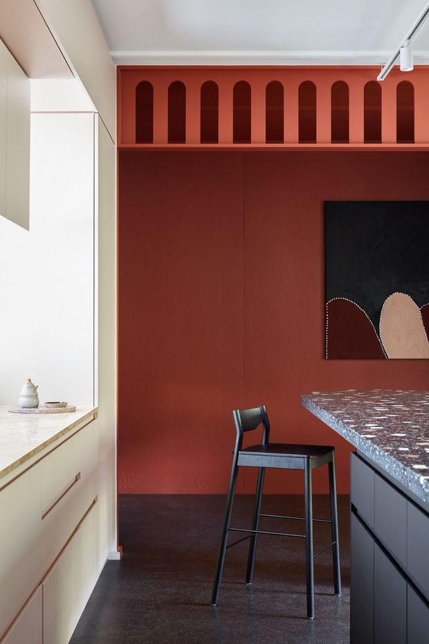 A compact, highly designed kitchen with a deep terracotta-red feature wall and matching arched display niches above a dark work area. Pale cabinetry and a light stone benchtop line the left side, while a dark island with a richly patterned stone surface occupies the right foreground. A black bar stool, track lighting, and abstract artwork complete the bold contemporary interior.