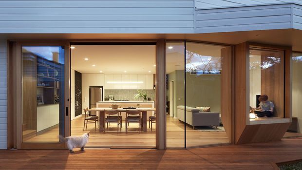 Contemporary residential extension viewed from a timber deck, with broad sliding glass doors opening to a bright combined kitchen and dining area. The interior features a long island, white cabinetry, timber dining furniture, and a grey sofa, while horizontal light-grey cladding and extensive glazing define the exterior.