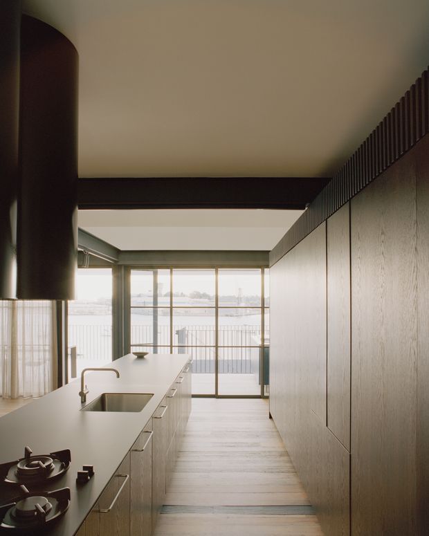 A narrow, contemporary kitchen with a long pale island and integrated sink, gas cooktop, light timber cabinetry, and a full-height bank of textured cabinetry along the right wall. Dark exposed ceiling beams and a large cylindrical rangehood create strong vertical and horizontal accents. Floor-to-ceiling glazing at the end of the room opens toward a balcony and bright waterfront or urban view.