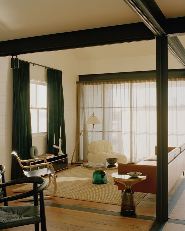 A warmly lit living room with a refined mid-century-inspired interior. The room features dark exposed structural beams and posts, expansive sheer-curtained glazing, deep green curtains, timber flooring, a textured area rug, sculptural lounge chairs, a cream upholstered sofa, and small organic-shaped side tables. The open glazed partition and strong black framing create a dramatic connection between adjoining spaces.