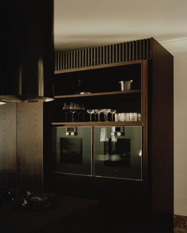 A dark, refined kitchen area featuring a tall built-in beverage and appliance cabinet in deep timber cabinetry. Open shelving displays wine glasses and tumblers, while two integrated glass-front ovens or appliances sit below. A black rangehood and dark benchtop are visible at left, with vertical slatted detailing along the cabinet top and light wall panelling in the background.
