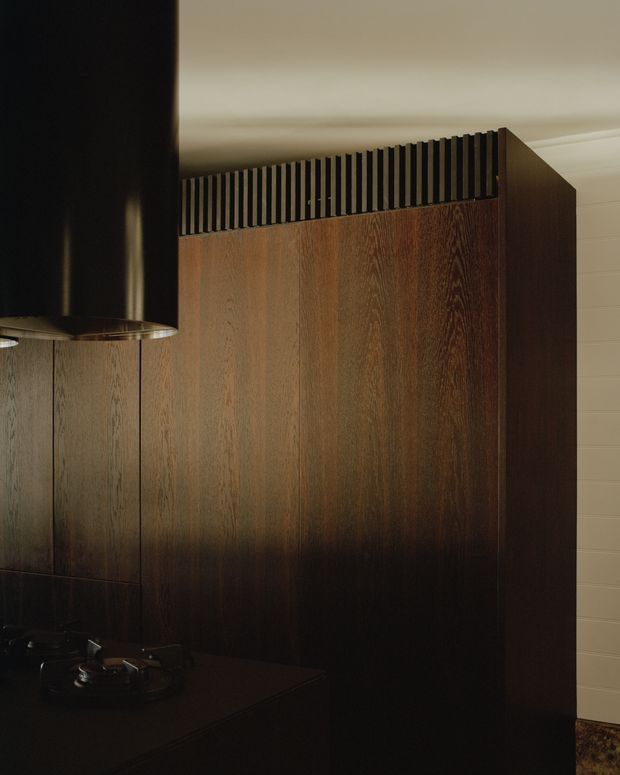 A moody close-up of a dark timber kitchen featuring tall vertically grained wood cabinetry, a black benchtop, and a large black cylindrical rangehood. A slatted timber ventilation detail tops the cabinetry, while pale horizontal wall panelling is visible at right.
