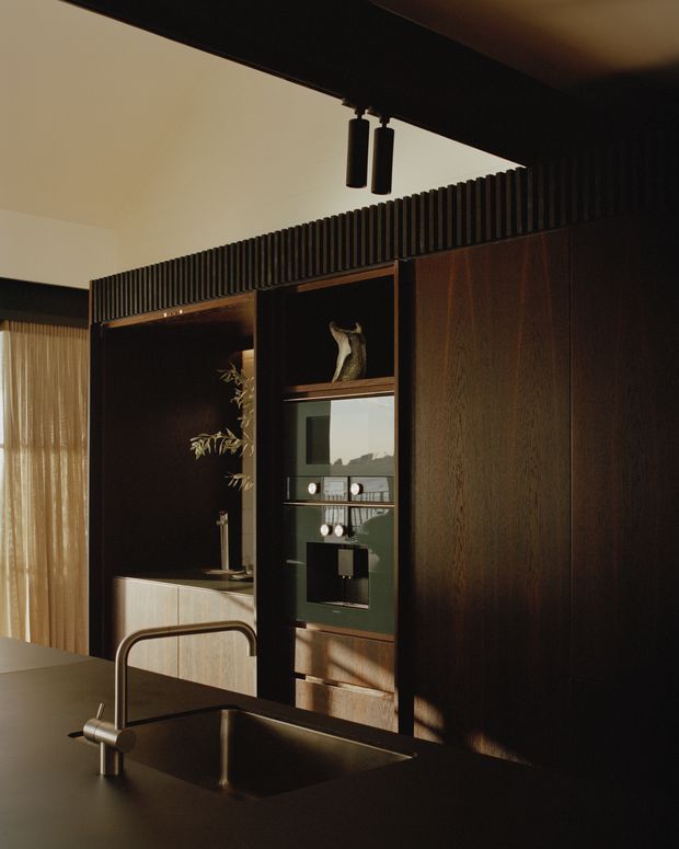Warm, moody interior view of a compact contemporary kitchen with dark timber-toned full-height cabinetry, a dark benchtop and integrated sink with brushed metal tapware. A ribbed overhead feature screen runs across the cabinetry, with integrated appliances and open display shelving. Soft daylight filters through sheer curtains at left, while a secondary sink or preparation area is visible in the background.