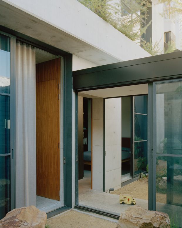 A modern courtyard-facing residential facade with a recessed timber entry door, dark green metal-framed glazing, exposed concrete slab projections, and a partially visible bedroom beyond the glazed opening. Natural rocks and planted greenery frame the entry, creating a compact indoor–outdoor connection.