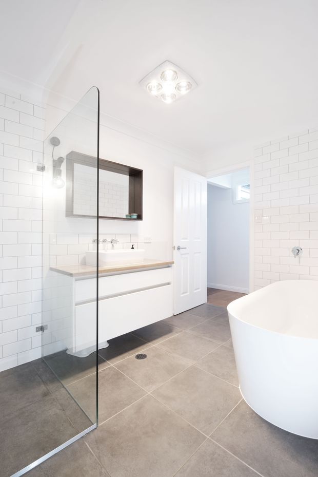 Bright contemporary bathroom featuring white subway-tile walls, a frameless glass shower screen, a floating white vanity with a light timber benchtop, a dark-framed recessed mirror, large grey floor tiles, and a freestanding white bathtub. A doorway leads to an adjacent space.