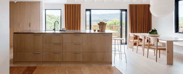 Bright open-plan interior featuring a warm timber kitchen with a large island, integrated cabinetry, black-framed windows, and a dining area beyond. Vertical timber screening accents, white walls and ceiling, light flooring, and expansive glazing create a contemporary, airy space with views to greenery and hills.