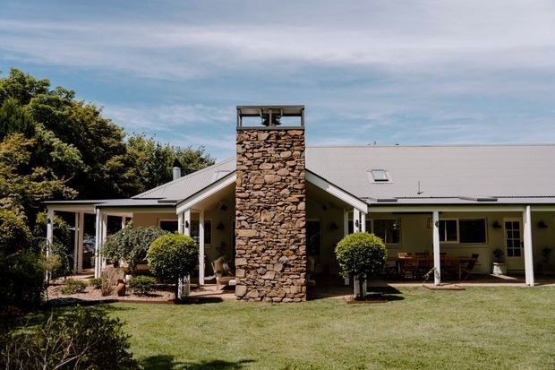 Single-storey residential house viewed from the front garden, with a broad low-pitched light metal roof, pale rendered walls, white-framed windows and verandah posts, and a prominent central rough-stone chimney extending above the roofline. The home is set within a landscaped suburban or rural garden with established trees and clipped shrubs.
