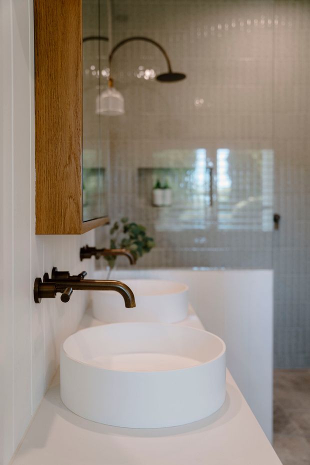 A warm, modern bathroom featuring two white vessel basins on a white vanity, wall-mounted bronze or aged brass faucets, a timber-framed mirrored cabinet, glossy light wall tiling, and a freestanding white bathtub visible in the background. A curved black-and-brass wall light and a small potted plant add understated decorative detail.
