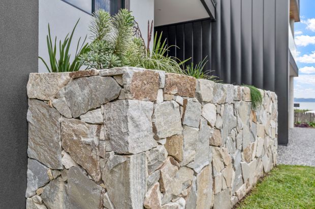 Close-up exterior view of a contemporary building with a substantial dry-stacked natural stone retaining or planter wall in the foreground. The wall contains drought-tolerant planting, while dark vertical cladding, smooth rendered surfaces, and a recessed entry or window area form the background. A gravel path and lawn are visible beside the building, with a bright distant landscape beyond.