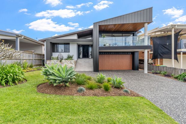 Front exterior view of a contemporary multi-level home with a distinctive sloping roof canopy, charcoal and light-grey facade, timber-lined upper balcony, glass balustrade, and a warm timber garage door. The property is approached by a gravel driveway and landscaped front garden featuring lawn, agave, and other low-maintenance planting.